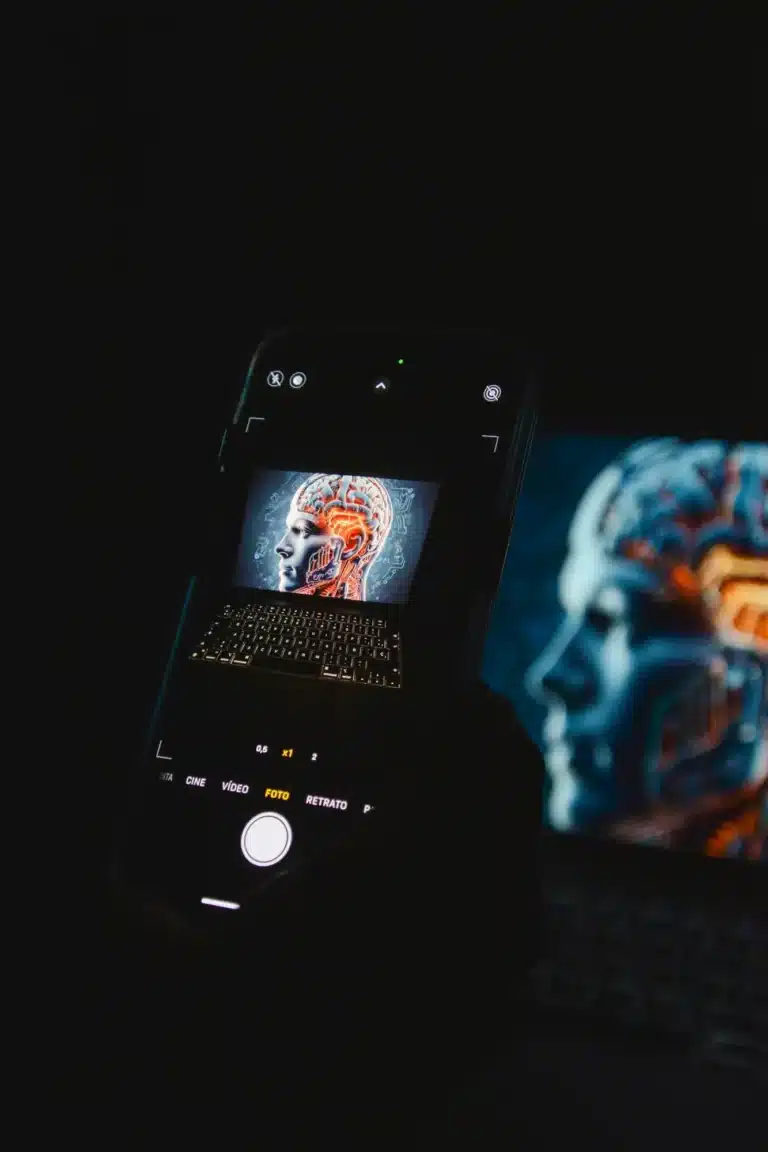 Dark scene with a smartphone showing a side profile of a head with an illuminated brain, while a blue-toned brain image glows on a nearby monitor.