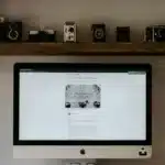 An iMac on a curved wooden stand sits on a desk, with vintage cameras lined on two shelves above.
