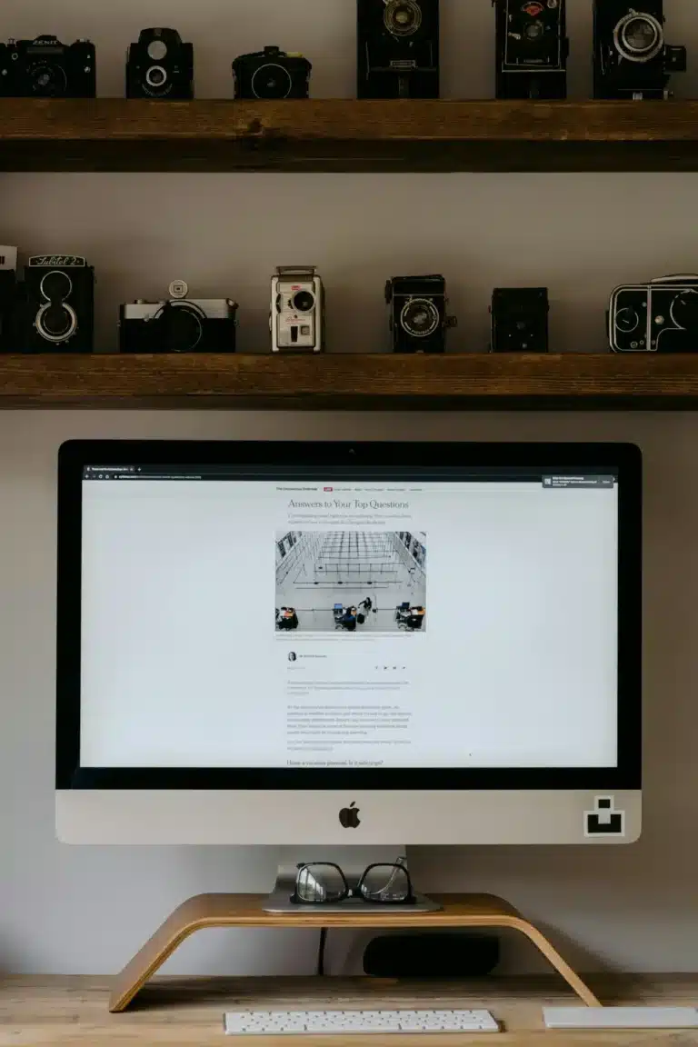 An iMac on a curved wooden stand sits on a desk, with vintage cameras lined on two shelves above.