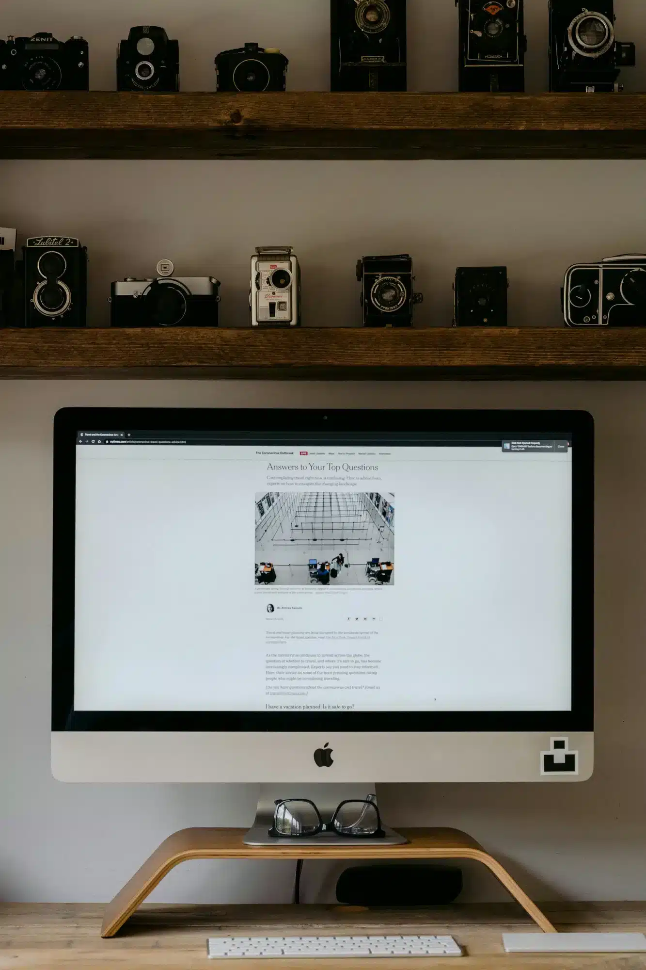 An iMac on a curved wooden stand sits on a desk, with vintage cameras lined on two shelves above.