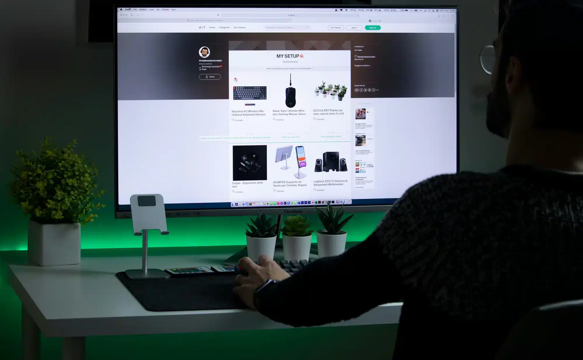 Person in a dark sweater sits at a white desk using a large monitor displaying the MY SETUP page, with small potted plants and green backlight.