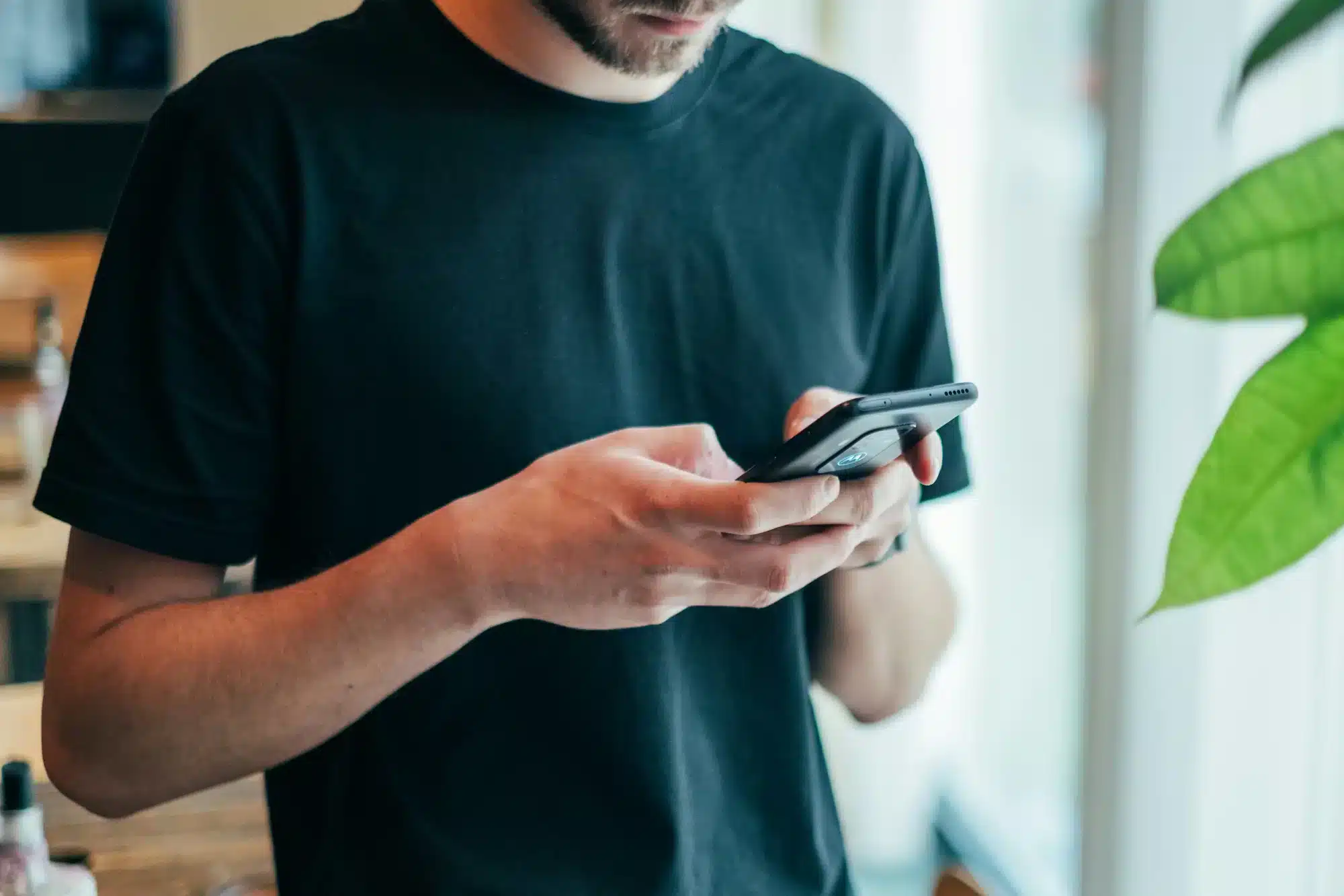 Man in a black T-shirt holding a smartphone indoors by a window with a green plant nearby.