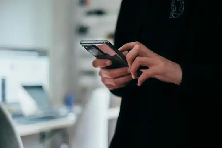 Close-up of hands holding two smartphones in a dim indoor office setting with a blurred desk in the background.