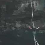 Person balances on a rope above dark water, arms outstretched, with rugged rock walls in the background.