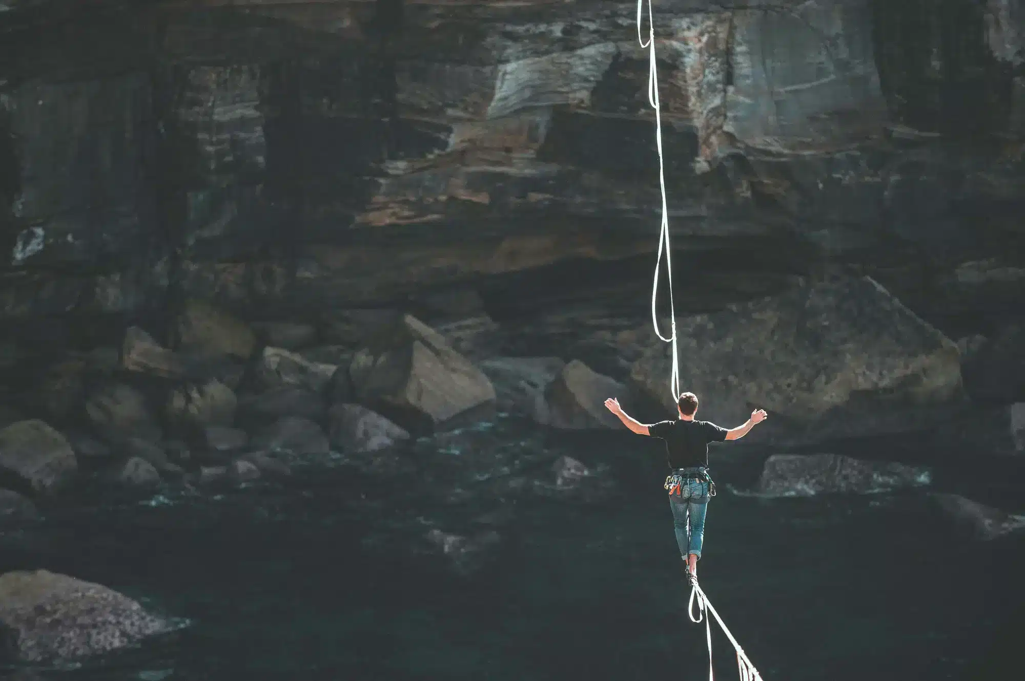 Person in a harness balances on a rope above dark water, arms outstretched, with rugged rock walls in the background.