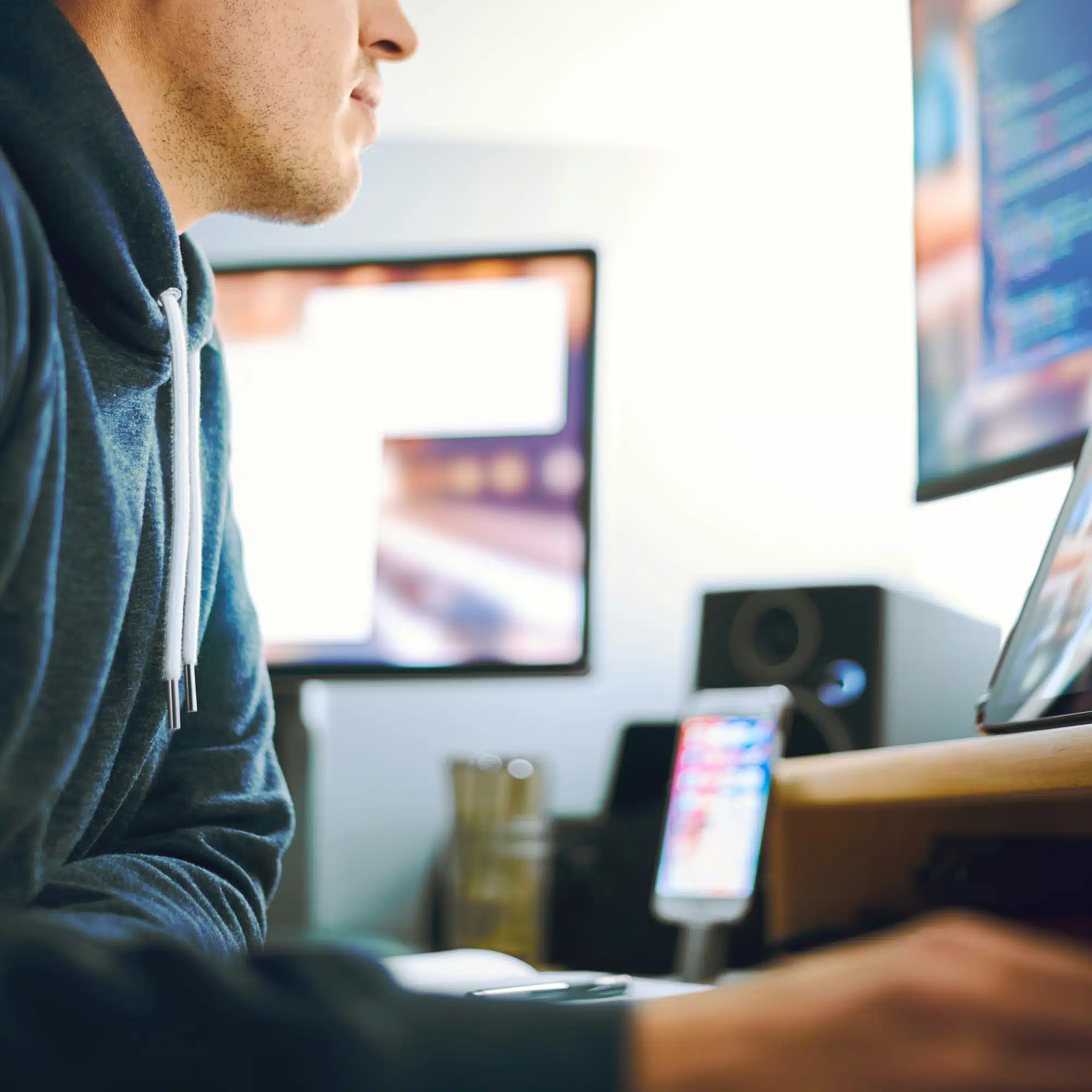 Side profile of a person wearing a dark hoodie, seated at a desk with multiple computer screens and a smartphone on a stand.