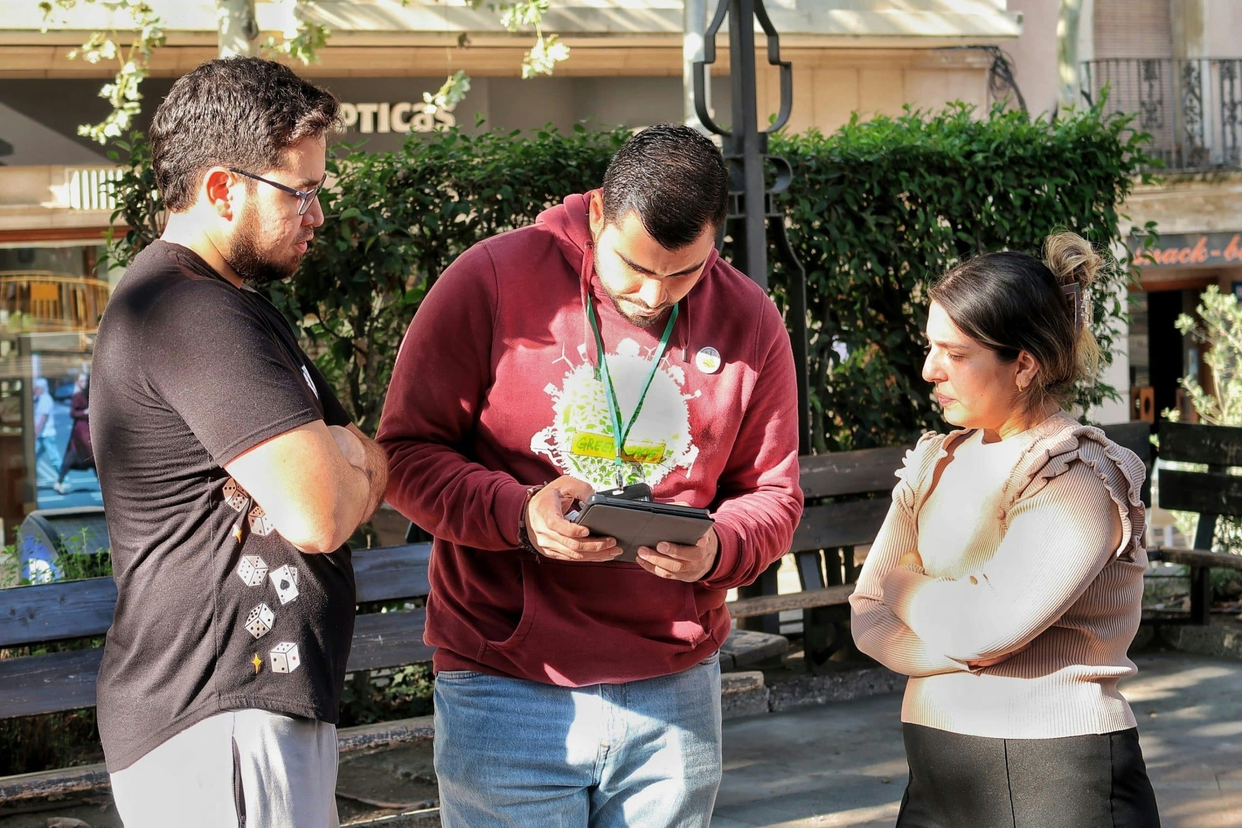 Three adults stand in a plaza, looking at a tablet: a man in a maroon hoodie with a green lanyard, a man in black, and a woman in pale pink.