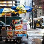 Street hot-dog cart under yellow and blue Sabrett umbrellas, with condiments, bottled drinks, and a hot-dog sign on a rainy city street.