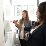 Two women in business attire discuss ideas at a glass whiteboard in a bright office, one writing with a red marker.
