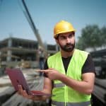Bearded construction worker in a neon vest and yellow hard hat holds a laptop and points at it on a busy construction site.