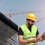 Construction worker in a neon safety vest and yellow hard hat examines a tablet at a construction site with scaffolding.