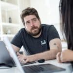 Bearded man in a dark gray t-shirt with a forearm tattoo sits at a white table with a laptop, looking at a person nearby in a bright office.