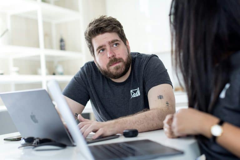 Bearded man in a dark gray t-shirt with a forearm tattoo sits at a white table with a laptop, looking at a person nearby in a bright office.