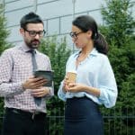 Man with glasses and a checkered shirt shows a tablet to a woman in a light blue blouse, outdoors, holding a coffee cup.