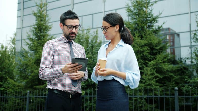 Man with glasses and a checkered shirt shows a tablet to a woman in a light blue blouse, outdoors, holding a coffee cup.