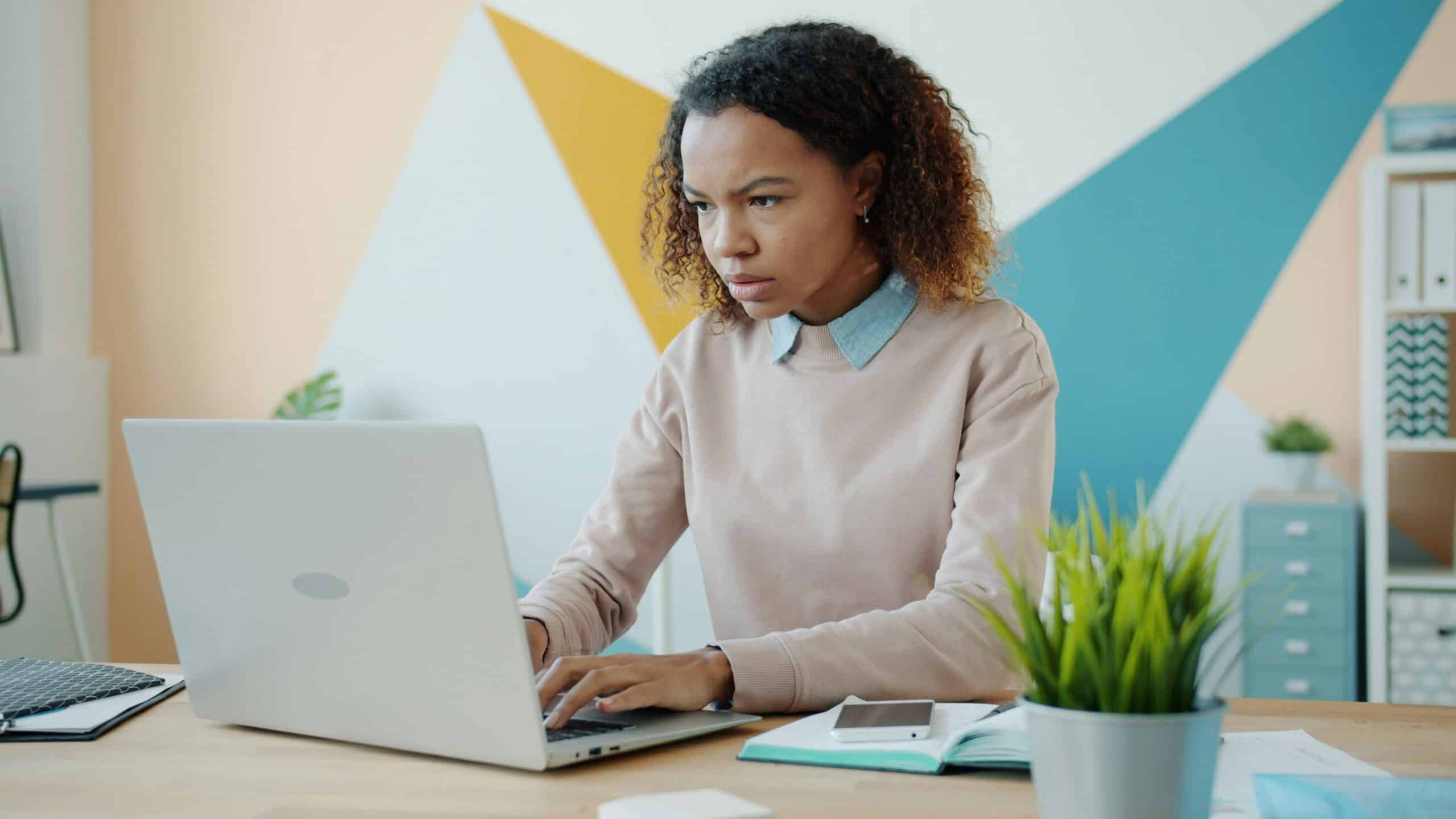 A focused woman types on a silver laptop at a desk in a bright office with geometric wall patterns and a potted plant.
