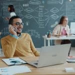 Man with glasses at a wooden desk talks on a cell phone, laptop open, papers nearby, in a modern office with chalkboard wall.