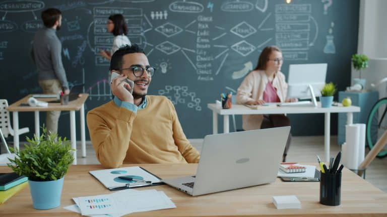 Man with glasses at a wooden desk talks on a cell phone, laptop open, papers nearby, in a modern office with chalkboard wall.