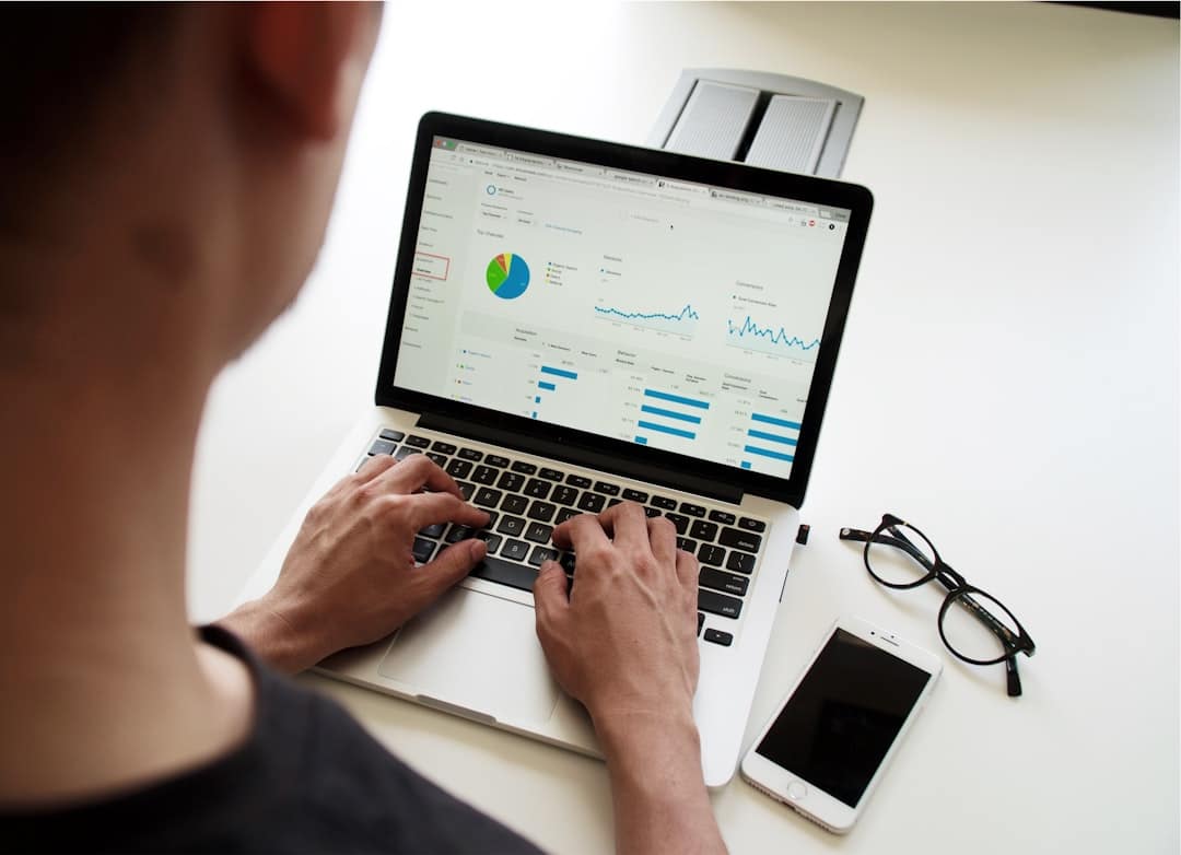 Person working on a laptop displaying analytics data, with a smartphone and glasses nearby. Photo by Campaign Creators on Unsplash.